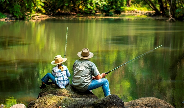young girl in hat with older man sitting on rocks in a coastal stream fishing 