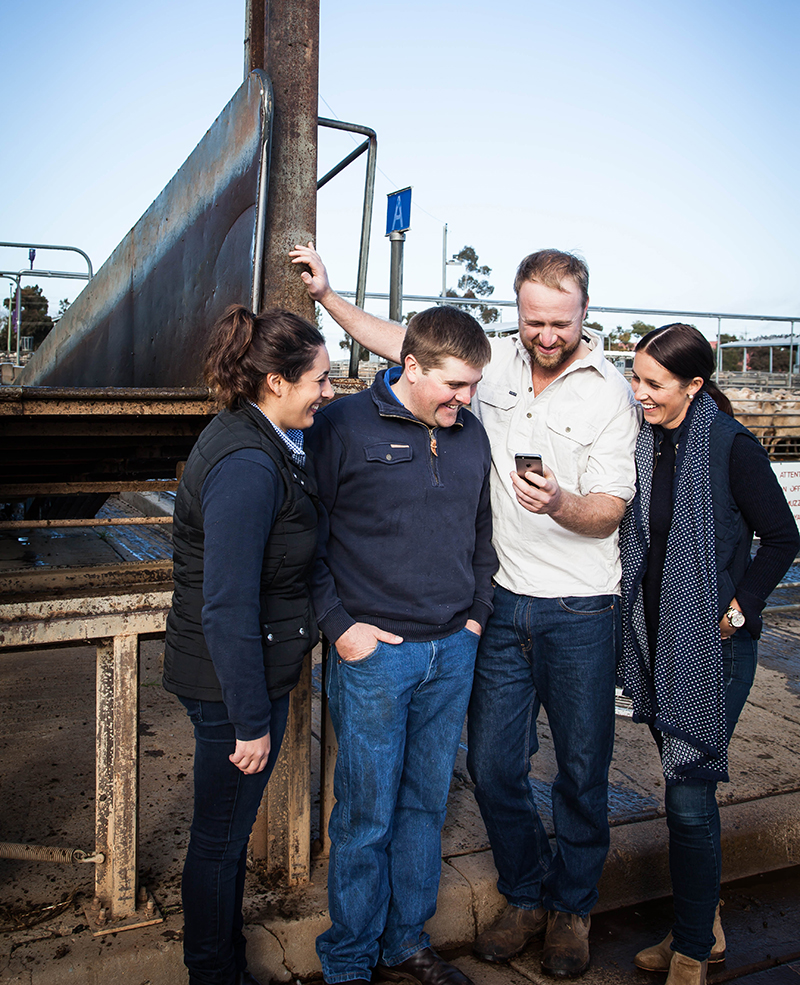 4 young farmers are smiling as they huddle around a mobile phone