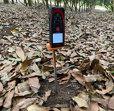 small sqaure laser metre inserted into ground, surrounded by leaves under tree canopy