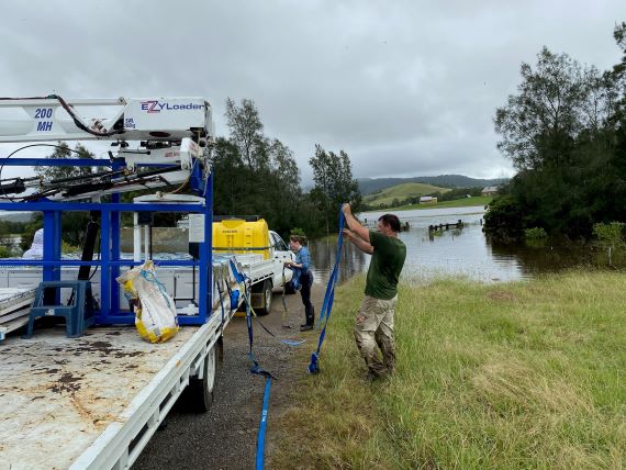 2 people loading beehives onto 2 utes in foreground, flood waters in background