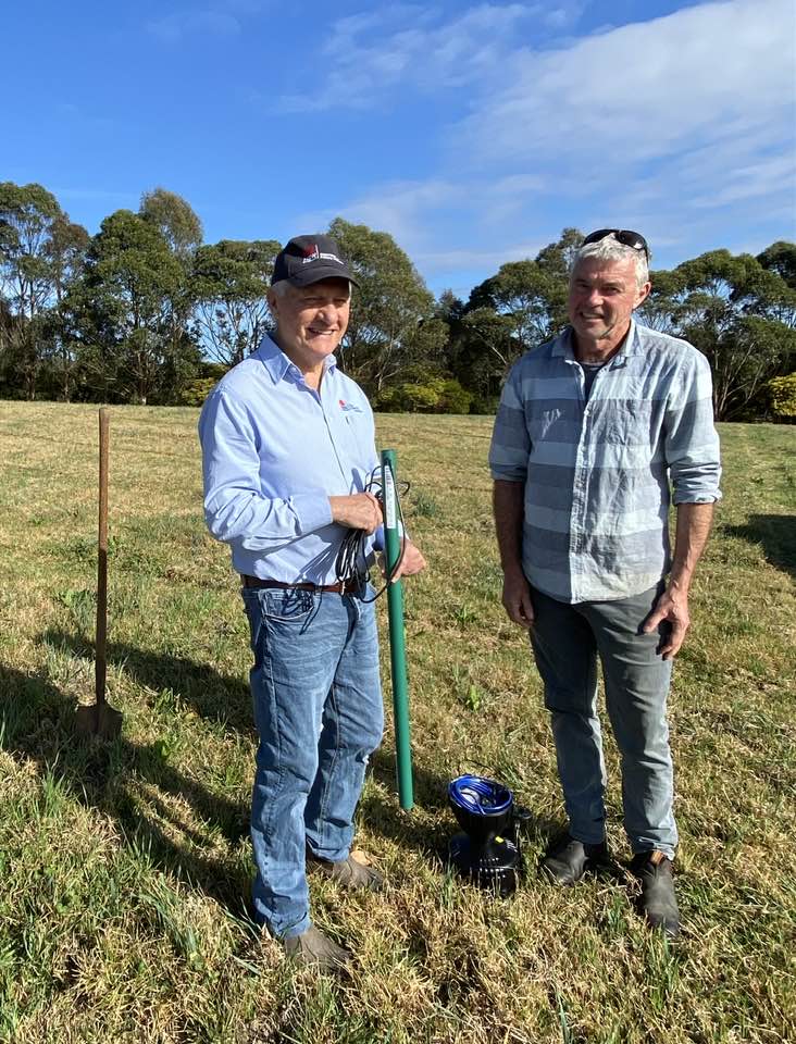 Luke Jewell on the left, wearing blue shirt and jeans holds long green monitoring device, and Dave O’Donnell in striped shirt and jeans, stand in a grassy paddock smiling for the camera.